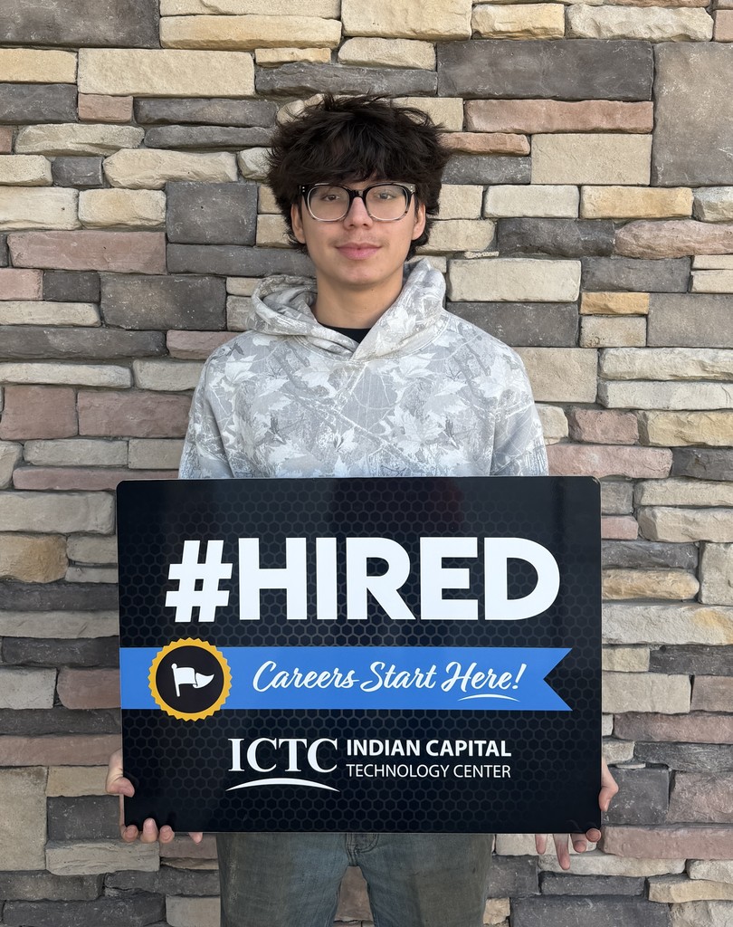 Person in glasses stands in front of brick wall, holding a black sign that says "#HIRED" and "Careers Start Here!"