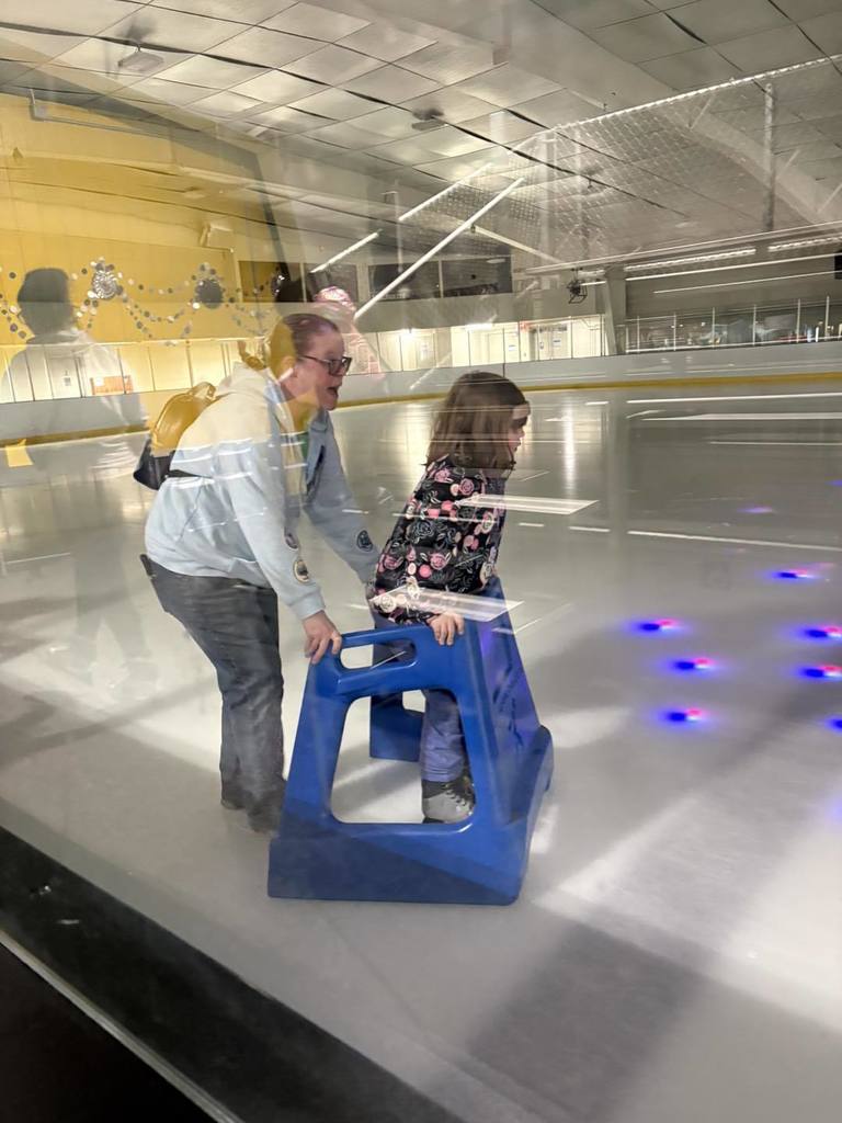 A student ice skating while holding on to a blue support cart in front of them and someone is helping them from behind. 
