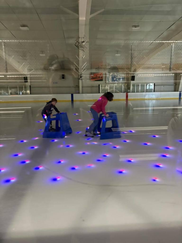 Students ice skating while holding on to a blue support cart in front of them. 