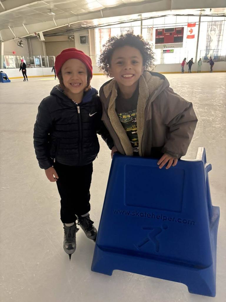 Students ice skating while one is holding on to a blue support cart in front of them. 
