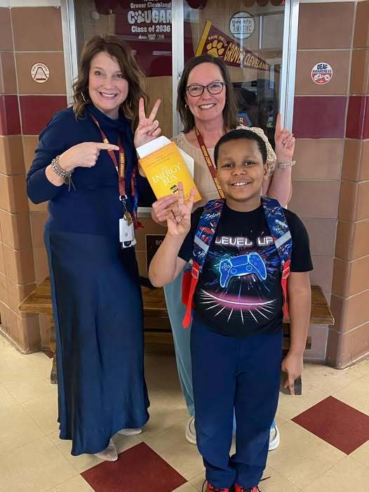 A student standing in front of two teachers and one teacher is holding a book.