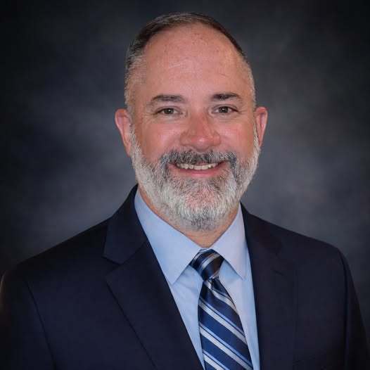 A man wearing a light blue dress shirt, a navy blue blazer with a striped tie is smiling. 
