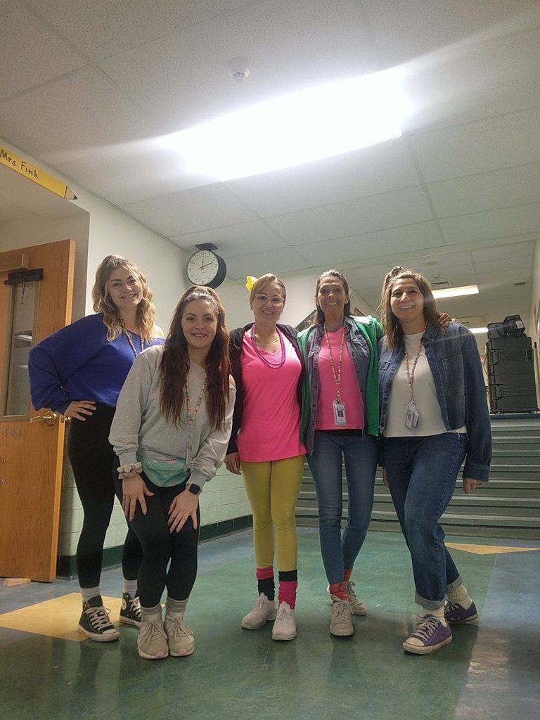 Teachers dressed like they are from the 80s and they are smiling in the hall. 