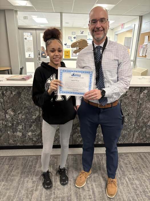 A young girl wearing a black hoodie and Principal Don Orlando holding the Stairclimber Award the student received.