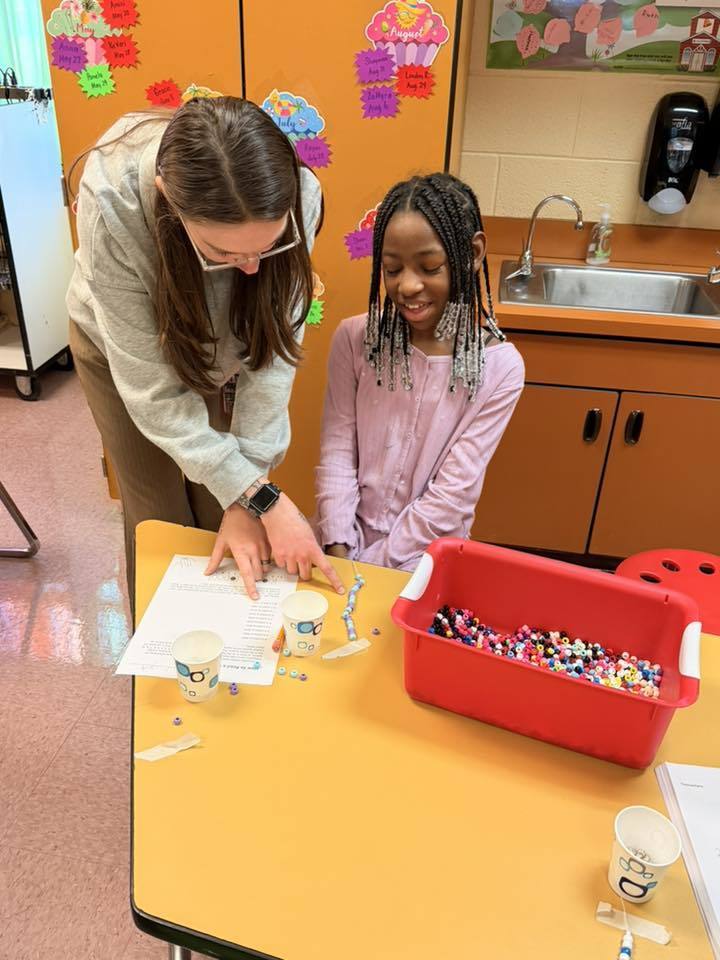 A teacher helping a student make a beaded bracelet. 