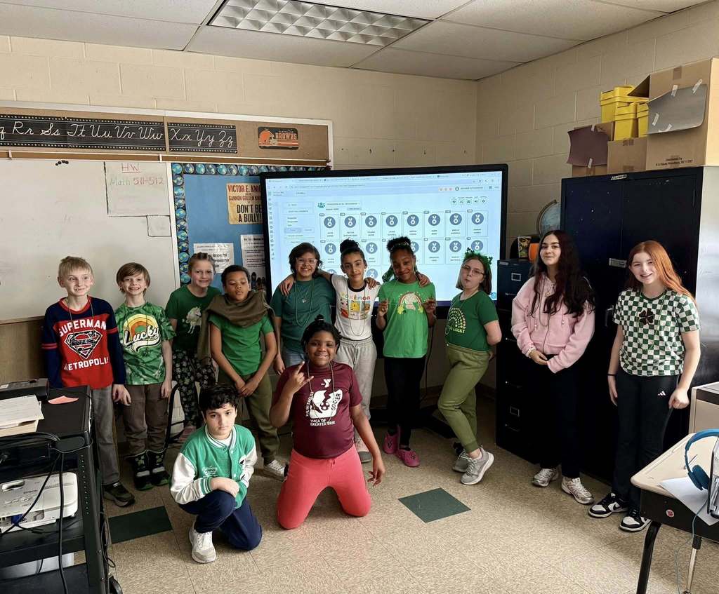 Students wearing green and standing in front of a large screen are smiling. 