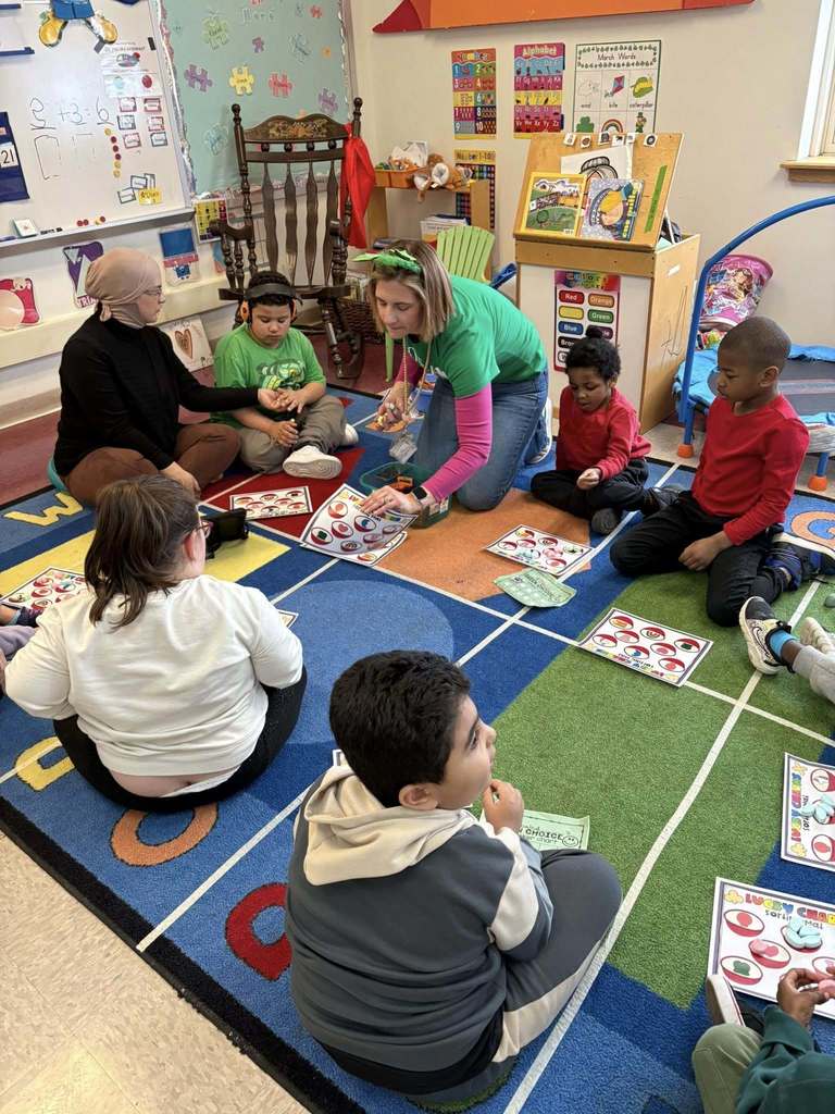 A teacher is wearing a green shirt and is sitting on a colorful rug while the students sort lucky charms on paper. 