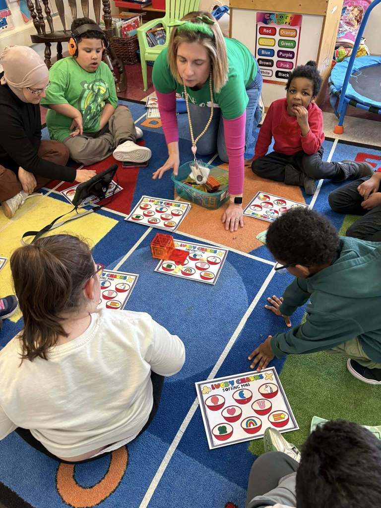 A teacher is wearing a green shirt and is sitting on a colorful rug while the students sort lucky charms on paper. 