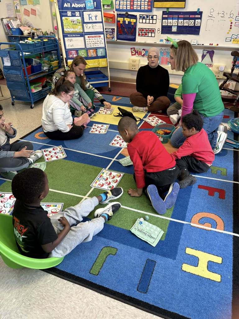 A teacher is wearing a green shirt and is sitting on a colorful rug with students. 