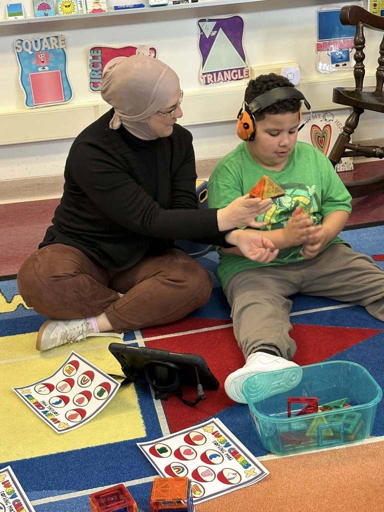 An adult is showing a pyramid she made to a student who is wearing headphones. 