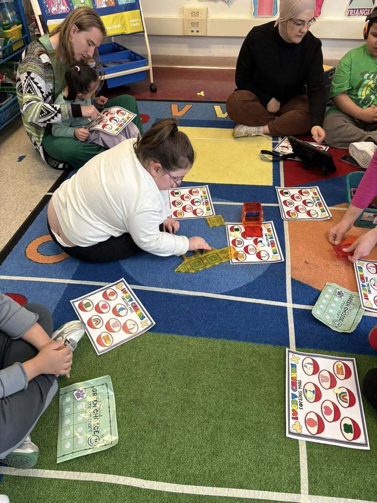 A teacher is wearing a green shirt and is sitting on a colorful rug while the students sort lucky charms on paper. 