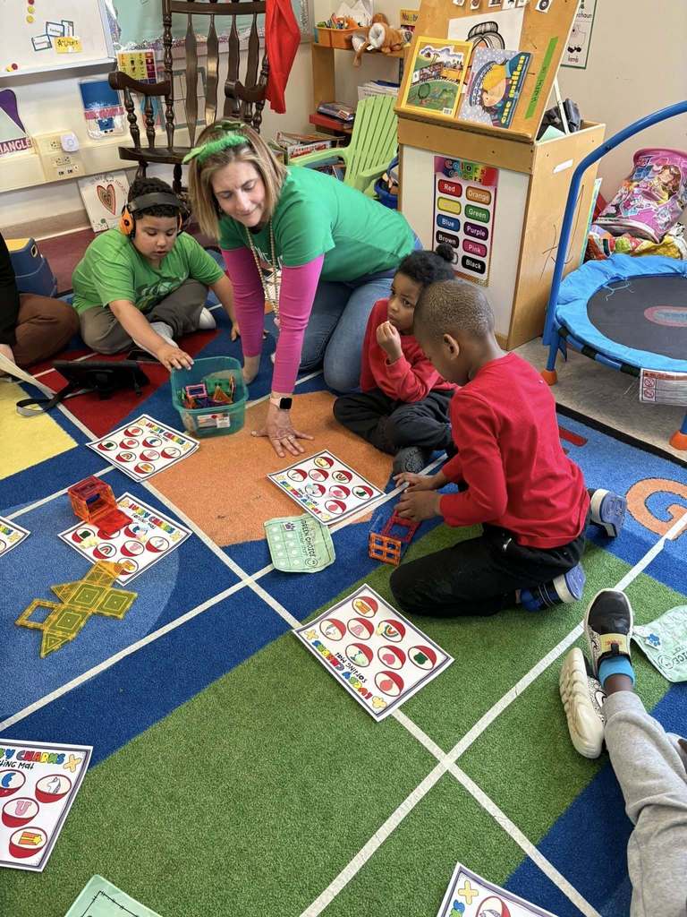A teacher is wearing a green shirt and is sitting on a colorful rug while the students sort lucky charms on paper.