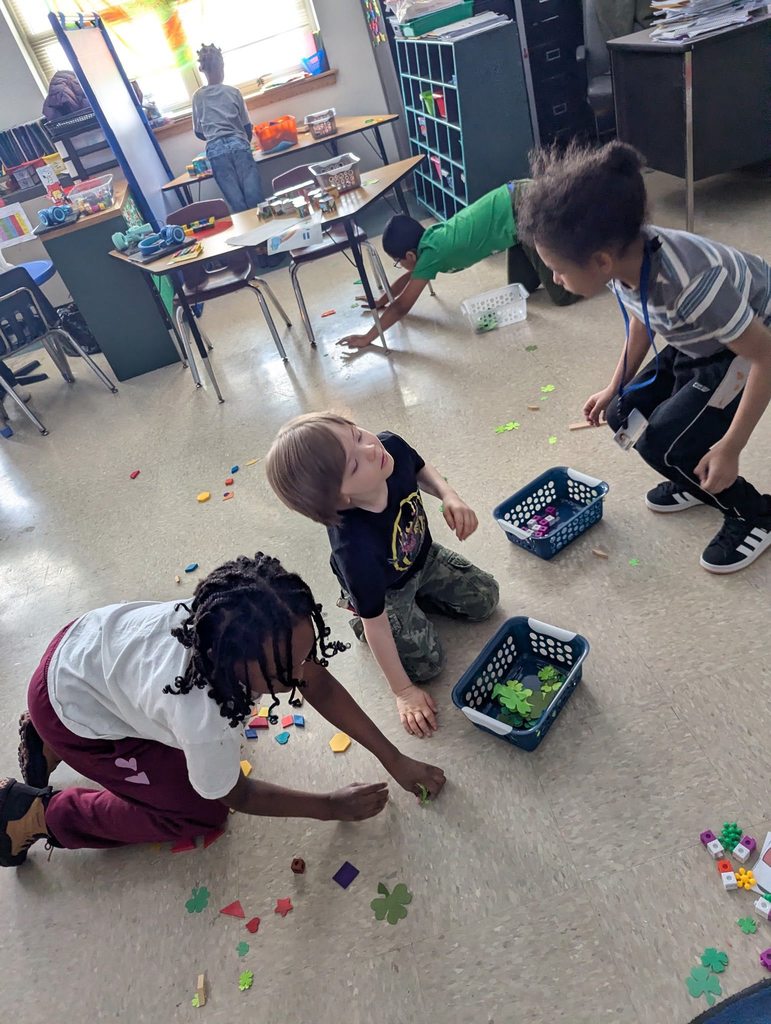 Students are picking up clovers off the floor in a classroom. 