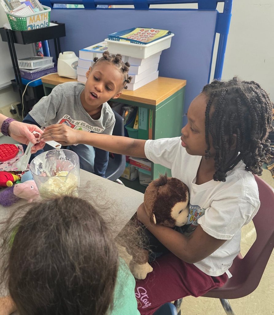 A student who is holding a stuffed monkey is helping put ingredients in to the blender to make a shamrock shake. 