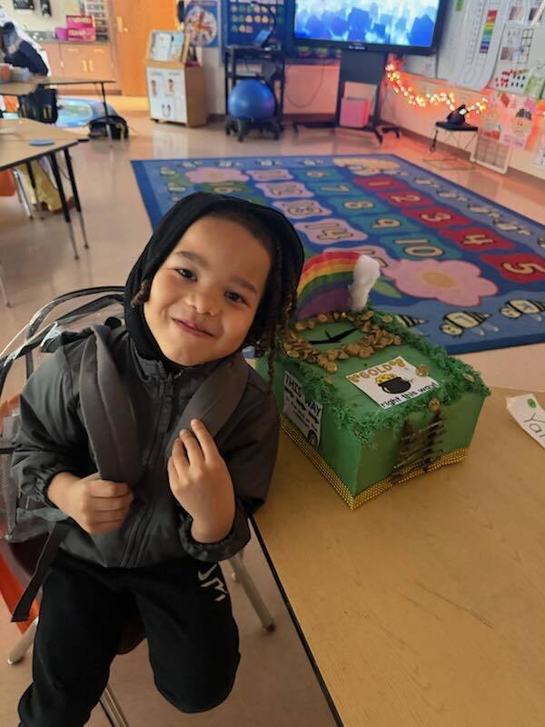 A student in a black jacket is smiling with his trap he made to try to catch a leprechaun on the table in front of him. 