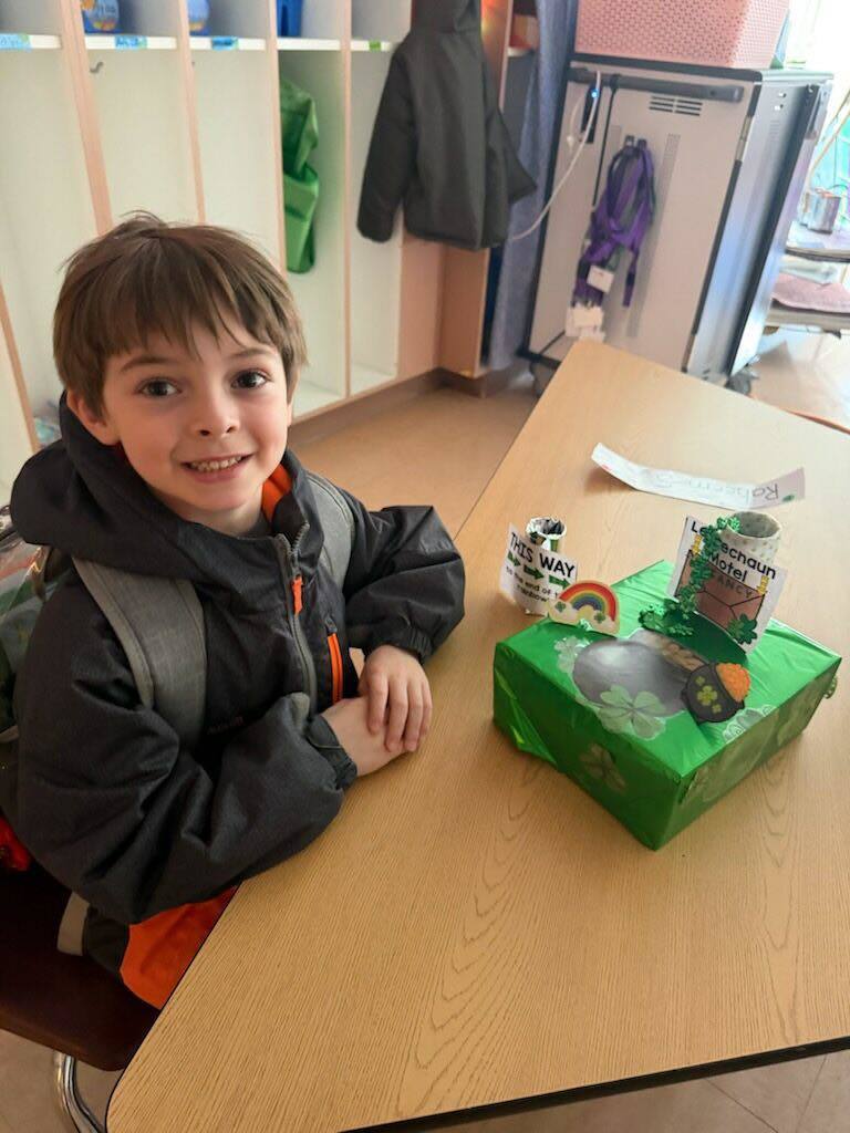 A student in a black jacket is smiling with his trap he made to try to catch a leprechaun on the table in front of him. 