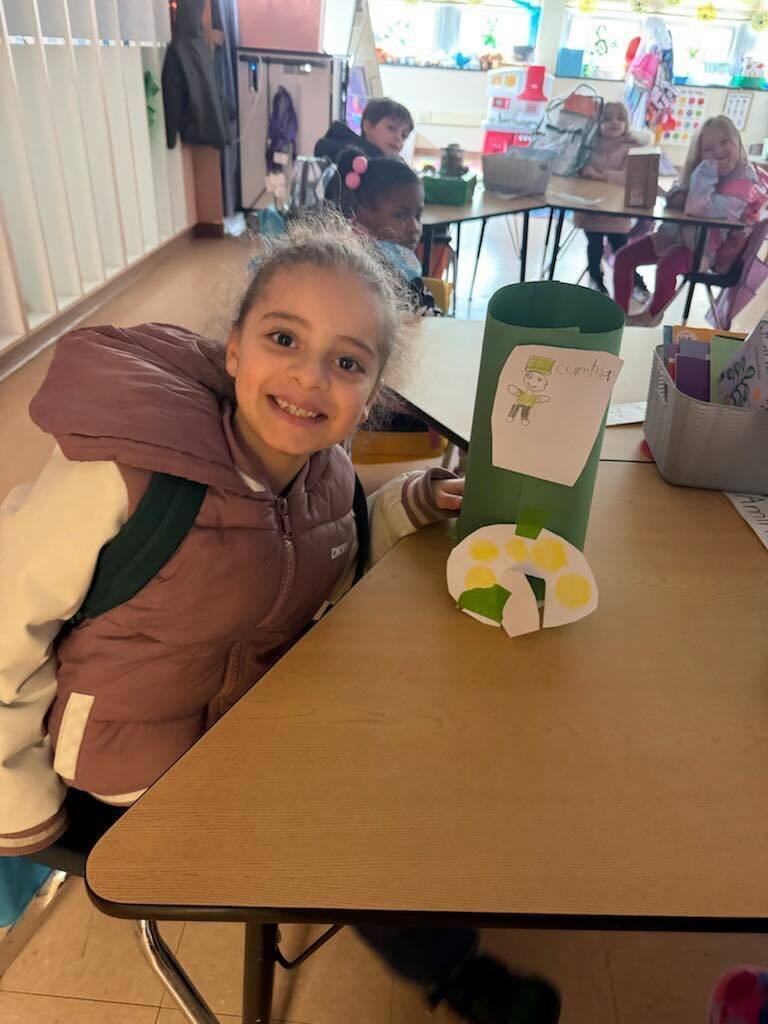A student in a purple and white jacket is smiling with her trap she made to try to catch a leprechaun on the table in front of her. 