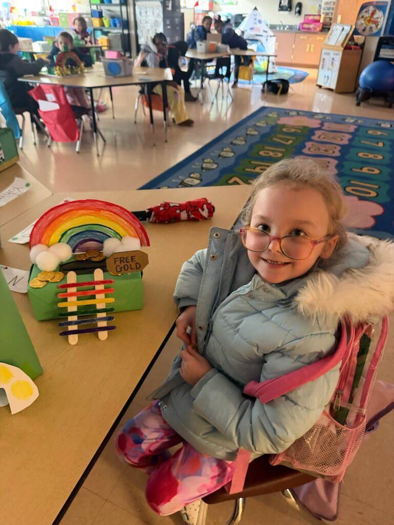 A student in a light blue jacket is smiling with her trap she made to try to catch a leprechaun on the table in front of her. 