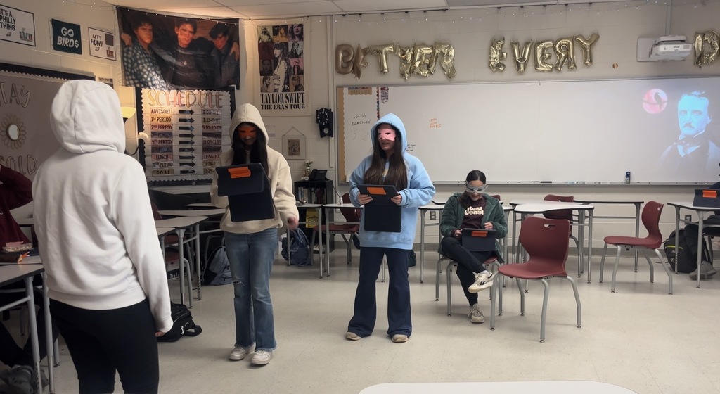 Four girls posed to recreate a scene in a short story. Classmates in desks looking on.