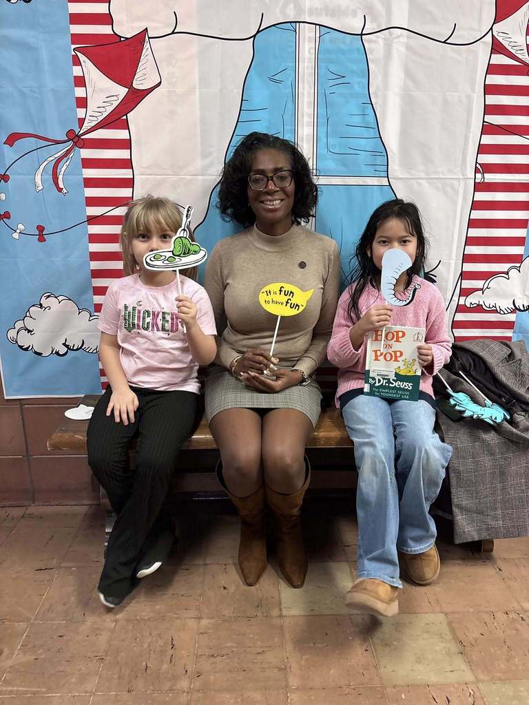 An adult and two students sitting on a bench holding a book and Dr. Suess props. 