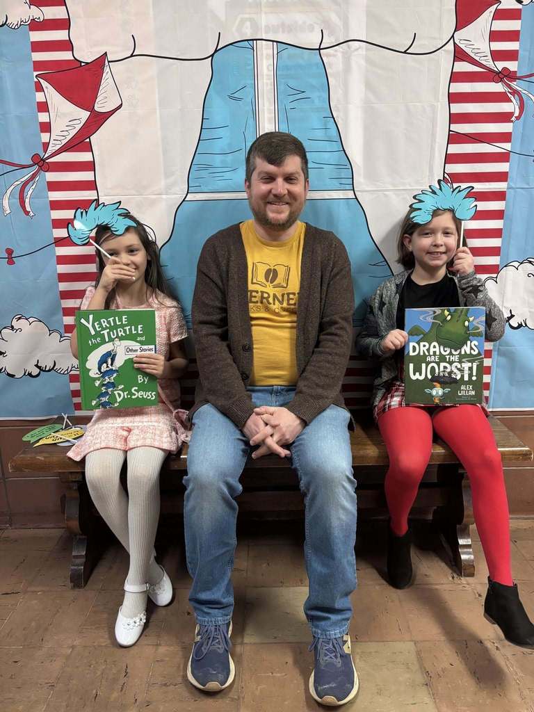 A man sitting with on a bench with students who are holding books and Dr. Suess props. 