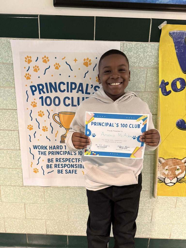 Young boy wearing a tan hoodieis holding his "Principal's 100 Club" Award. 