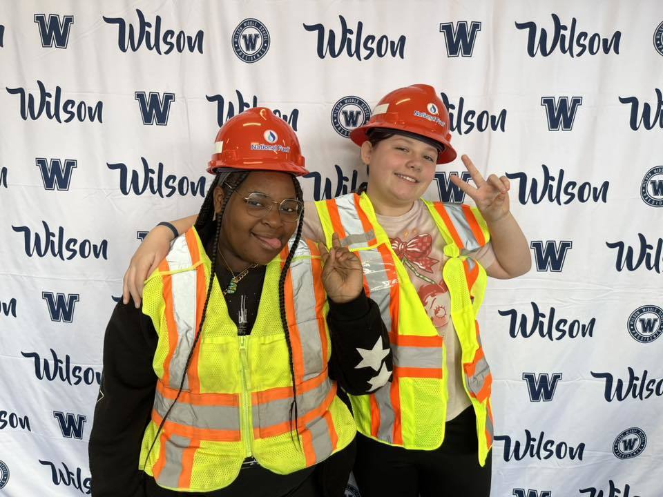 Students wearing National Fuel hard hats and vests while they smile and hold up peace signs. 