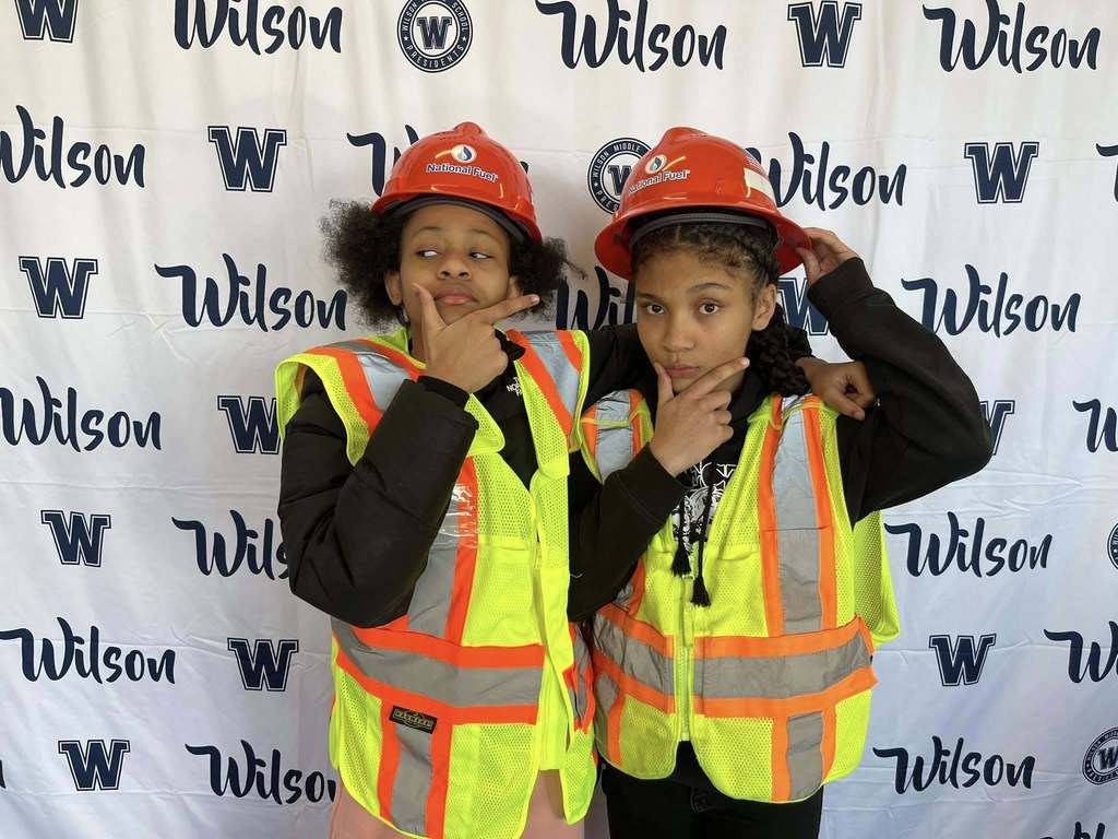 Students wearing National Duel hard hats and vests while they smile and holding two fingers on their chins as if they are thinking. 
