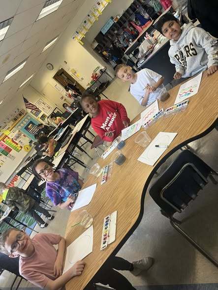 Students sitting at a table with paper and paint in front of them. 