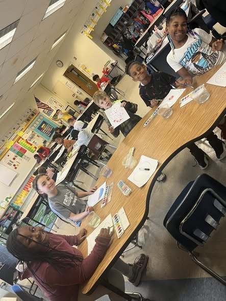 Students sitting at a table with paper and paint in front of them. 