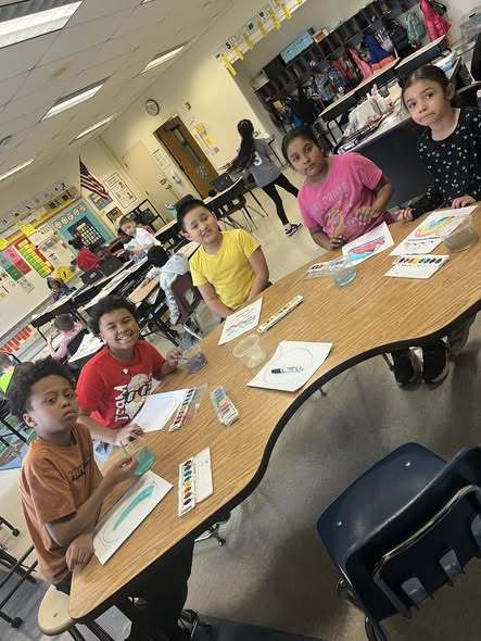 Students sitting at a table with paper and paint in front of them. 