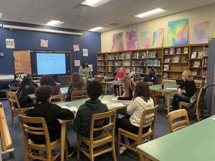 A women speaking to students who are sitting at tables in a library. 