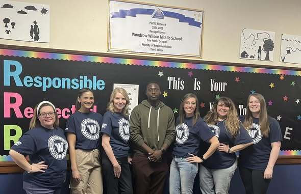 Staff wearing matching blue shirts that have the Wilson Middle School logo on them and a young man who is wearing a green hoodie is standing in the middle. 