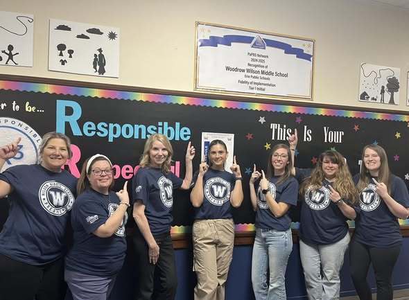 Staff wearing matching blue shirts that have the Wilson Middle School logo on them and they are pointing up. 