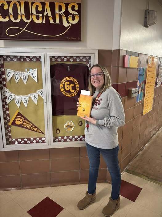A teacher wearing a gray shirt and jeans is holding a yellow book. 