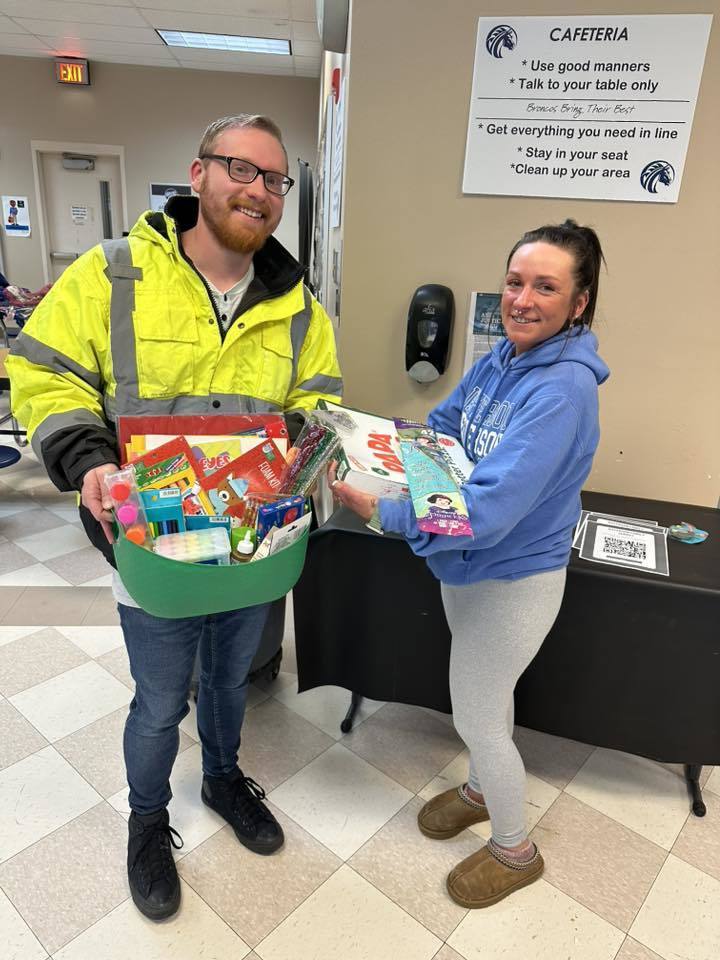 A man in a bright yellow jacket is holding a basket of goodies while a women next to him in a blue hoodie is holding games and other goodies in her hands. 