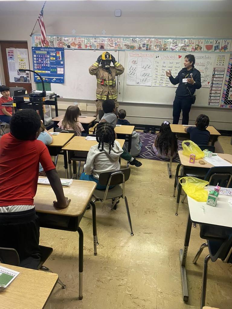 A firefighter dressed up in his gear in a classroom, while a man talks to the class. 
