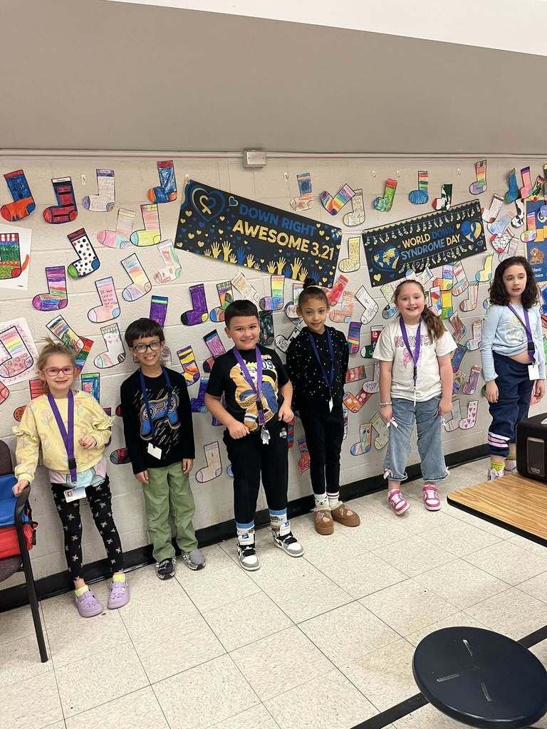 Students standing in front of the colorful cut outs of socks that students colored are hanging on the wall. 