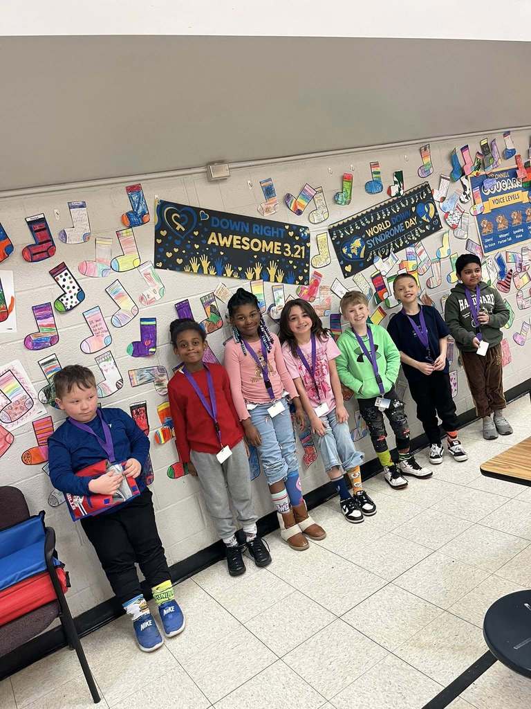 Students standing in front of the colorful cut outs of socks that students colored are hanging on the wall. 