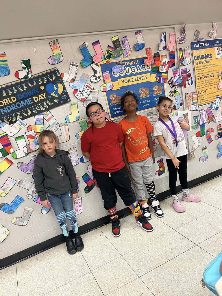 Students standing in front of the colorful cut outs of socks that students colored are hanging on the wall. 