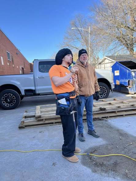 A young man in an orange shirt is talking to a man in a brown jacket. 