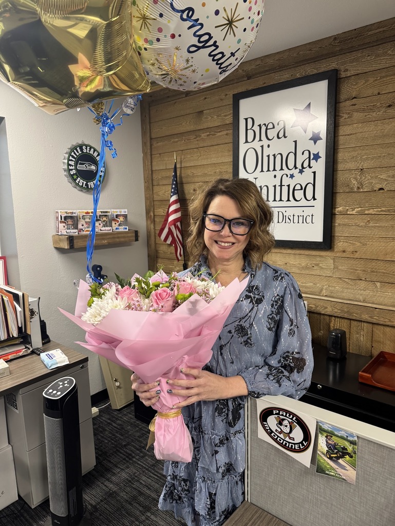 Classified employee of the year Jamie Robinson holds a pink bouquet of flowers and gold and white balloons. She is in front of a large sign with the BOUSD logo