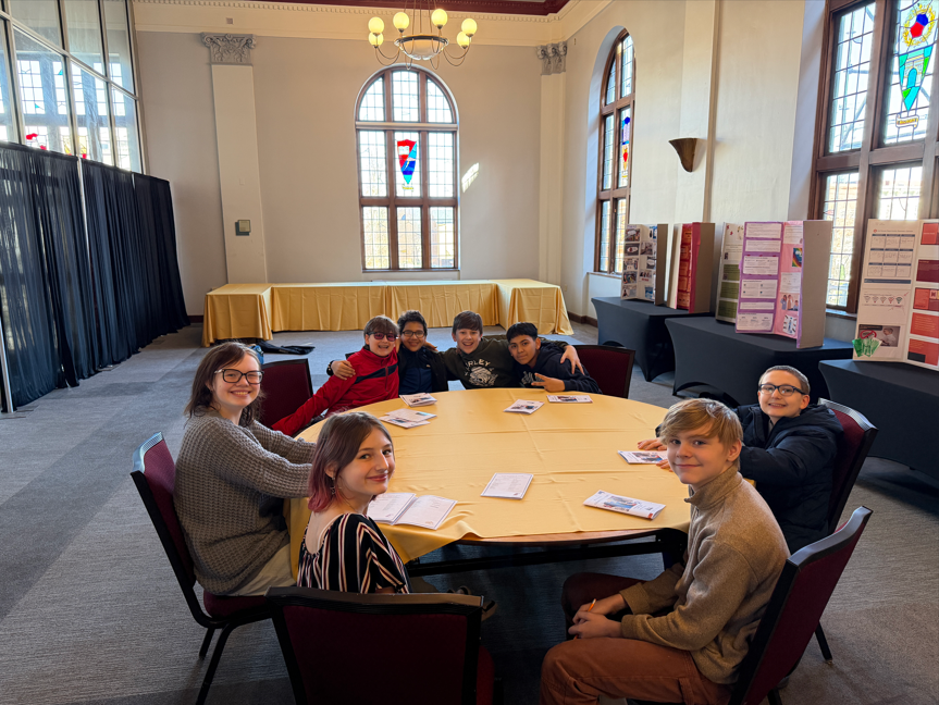 A group of students sitting at a circular table. and a hidden easter egg is on the table behind them. 