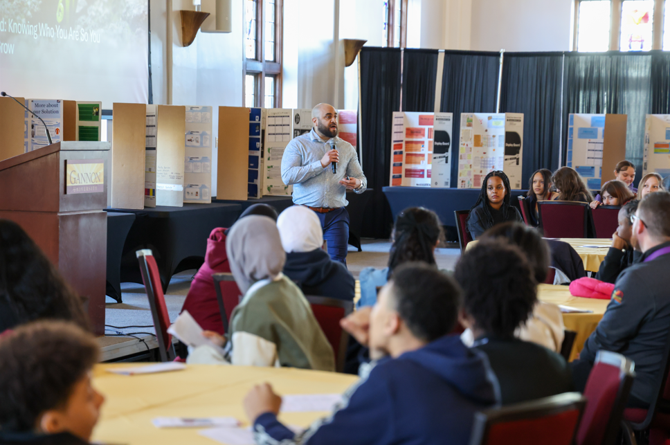 A man in a light blue dress shirt is holding a microphone and is speaking to a room full of kids sitting at tables.