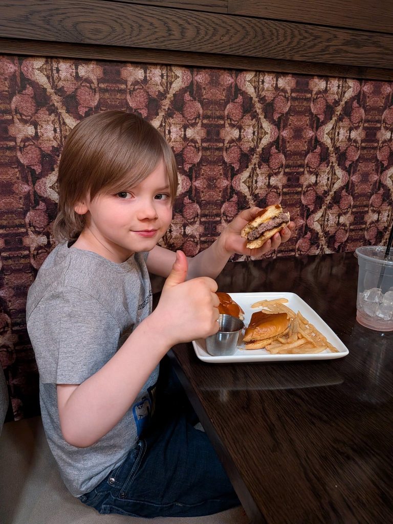 A young kid wearing a gray shirt is holding a hamburger in one hand and giving a thumbs up with his other hand. 