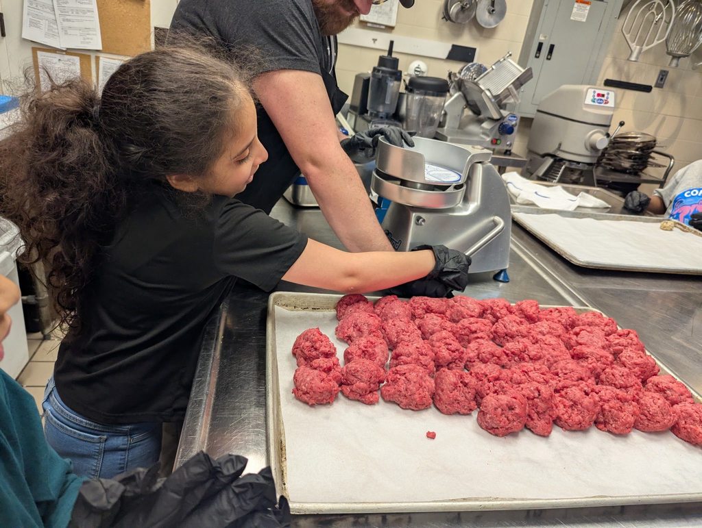 A student wearing a black shirt is helping press meat to make it into a patty for burgers. 