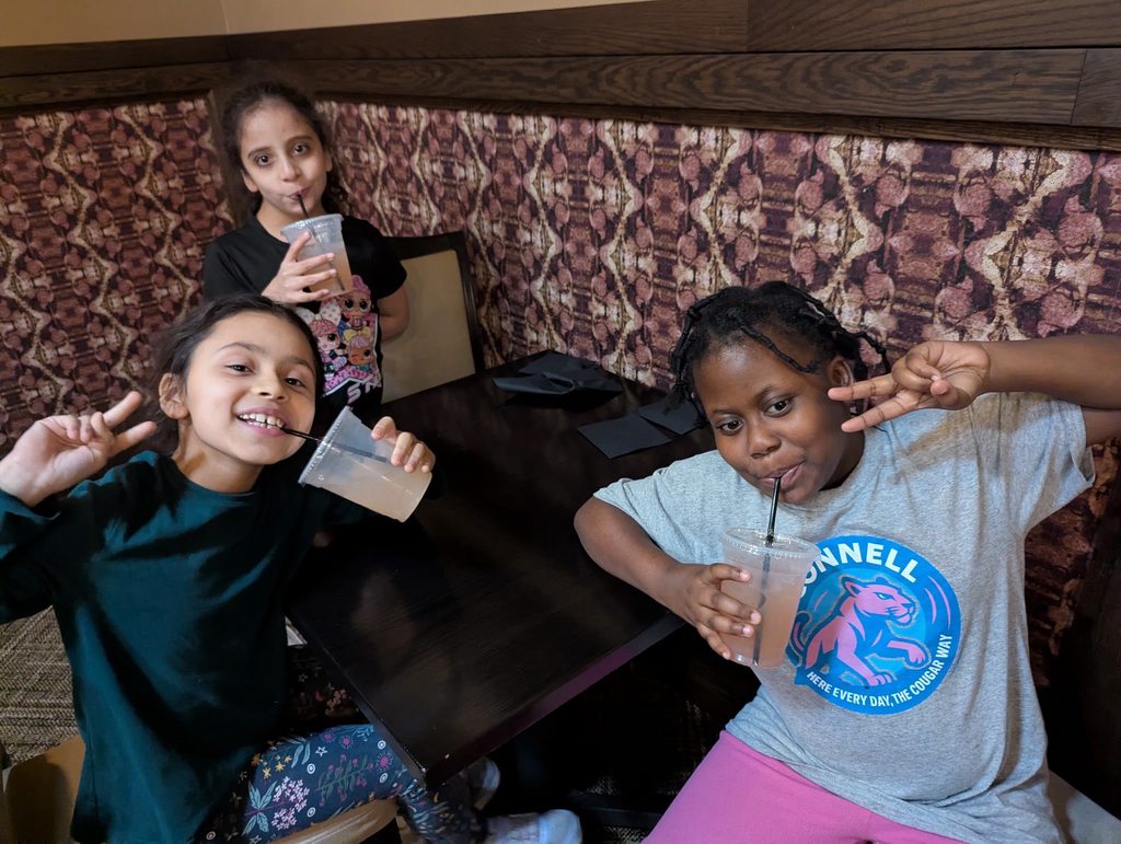 Three students sitting at a table and drinking a juice from straws while two of them hold up peace signs. 