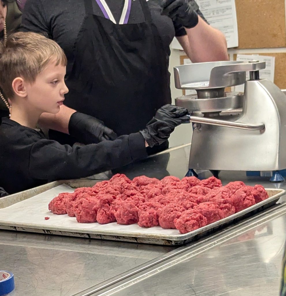 A student wearing a black shirt is helping press meat to make it into a patty for burgers. 
