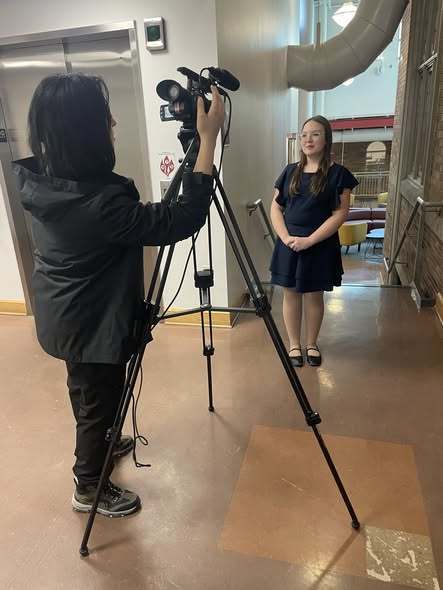 A student in a navy blue dress is getting interviewed by a women holding a camera. 