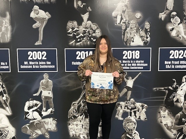 EAJ Student of the Week Aaliyah Keene holding her certificate in front of the Mt. Morris Athletic mural.